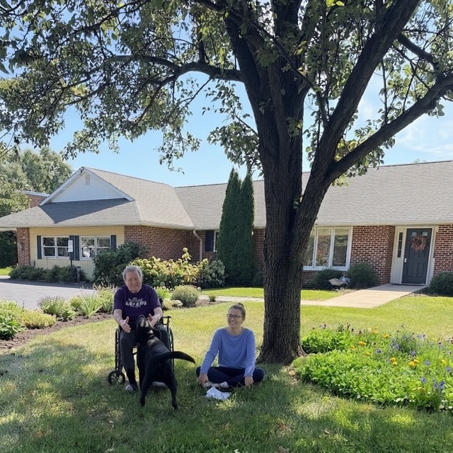 Resident in wheelchair enjoying time outdoors with a visitor and dog at Madonna Heritage